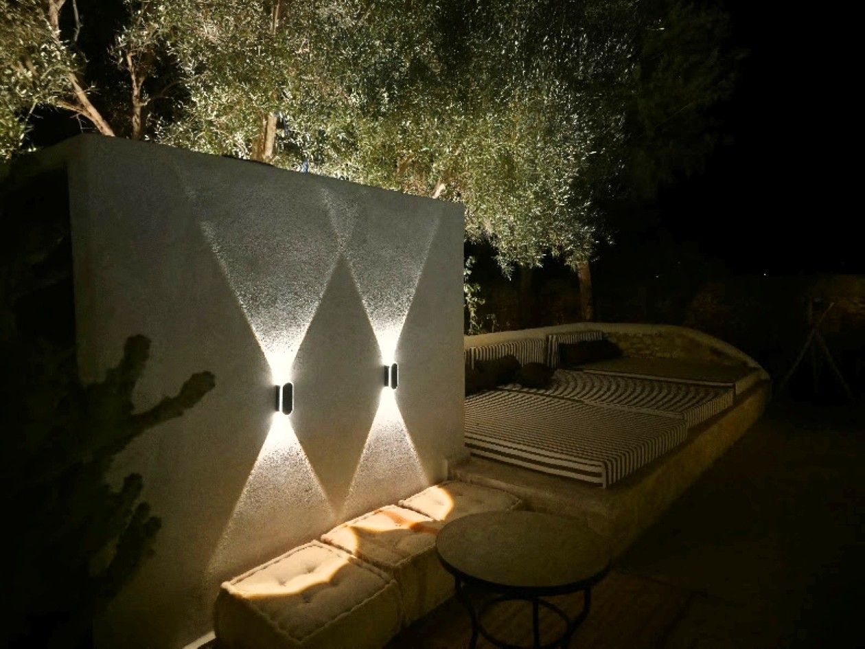 An outdoor seating area with a curved bench and a small table, adjacent to a wall with two illuminated wall sconces.