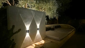 An outdoor seating area with a curved bench and a small table, adjacent to a wall with two illuminated wall sconces.