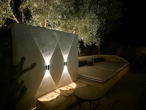 An outdoor seating area with a curved bench and a small table, adjacent to a wall with two illuminated wall sconces.