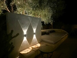 Un área de estar al aire libre con un banco curvo y una mesita pequeña, junto a una pared con dos apliques de luz.