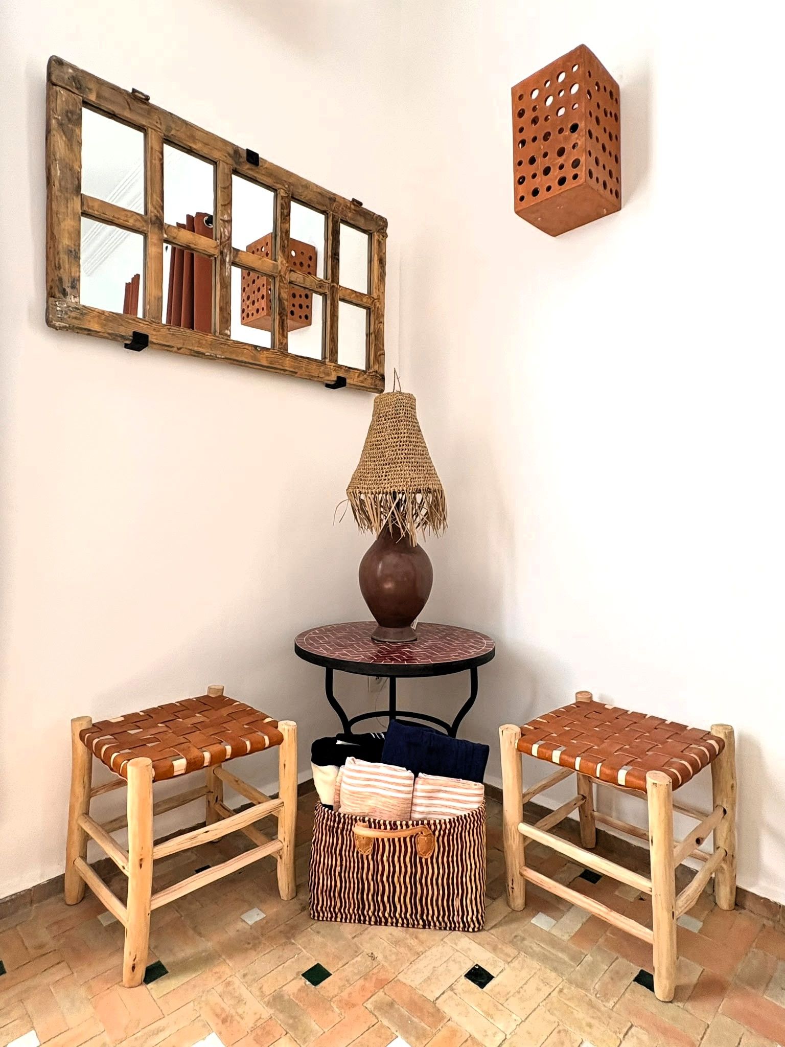 A corner room with a rustic wooden mirror, a woven lamp, two woven chairs, a small round table, and a woven basket.
