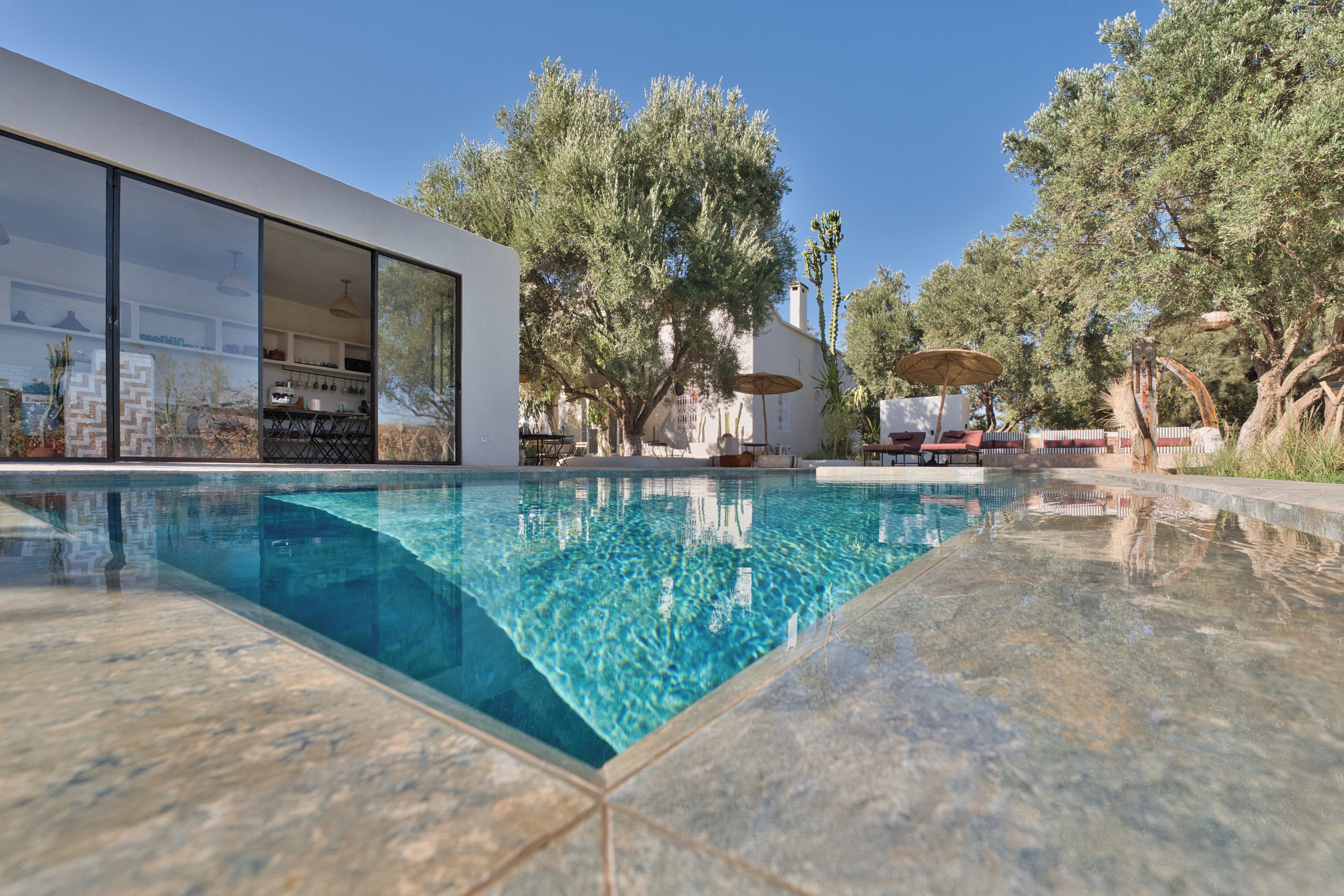 Una piscina moderna al aire libre con agua azul cristalina, rodeada por un patio con mobiliario exterior y un paraguas grande.
