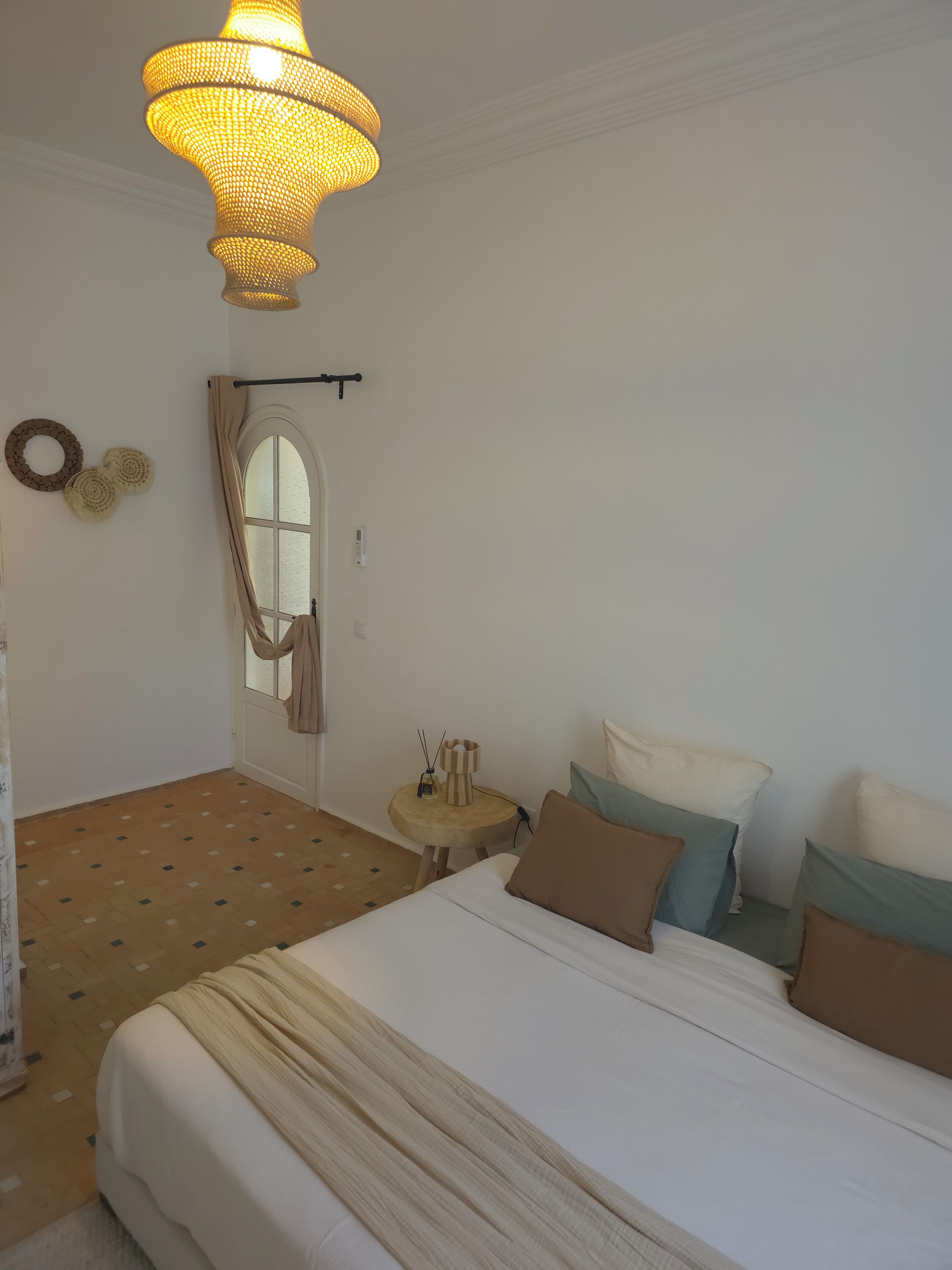 A bedroom with a large bed, a bedside table with a lamp, a hanging light fixture, and a window with curtains.