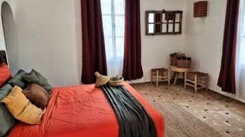 A bedroom with a bed, a window with curtains, a small table, and a chair.