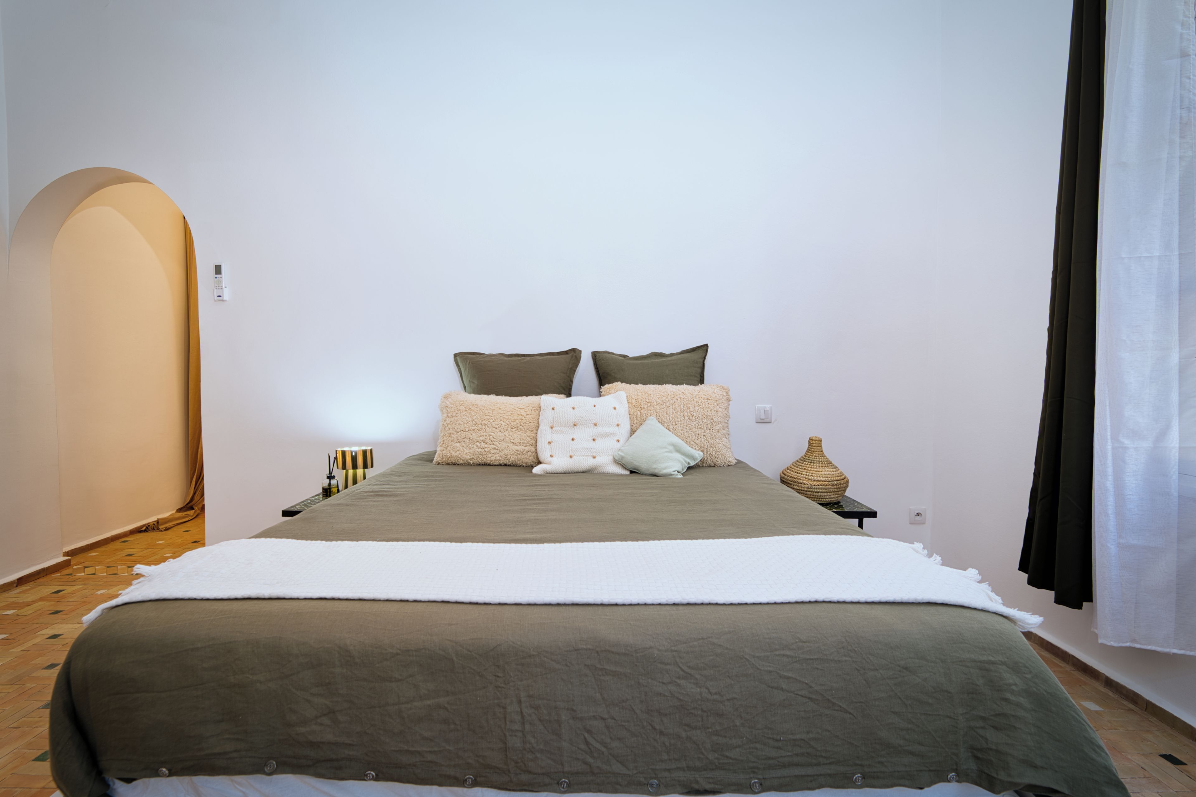 A bedroom with a bed, pillows, a bedside table, a lamp, and a wall-mounted switch.