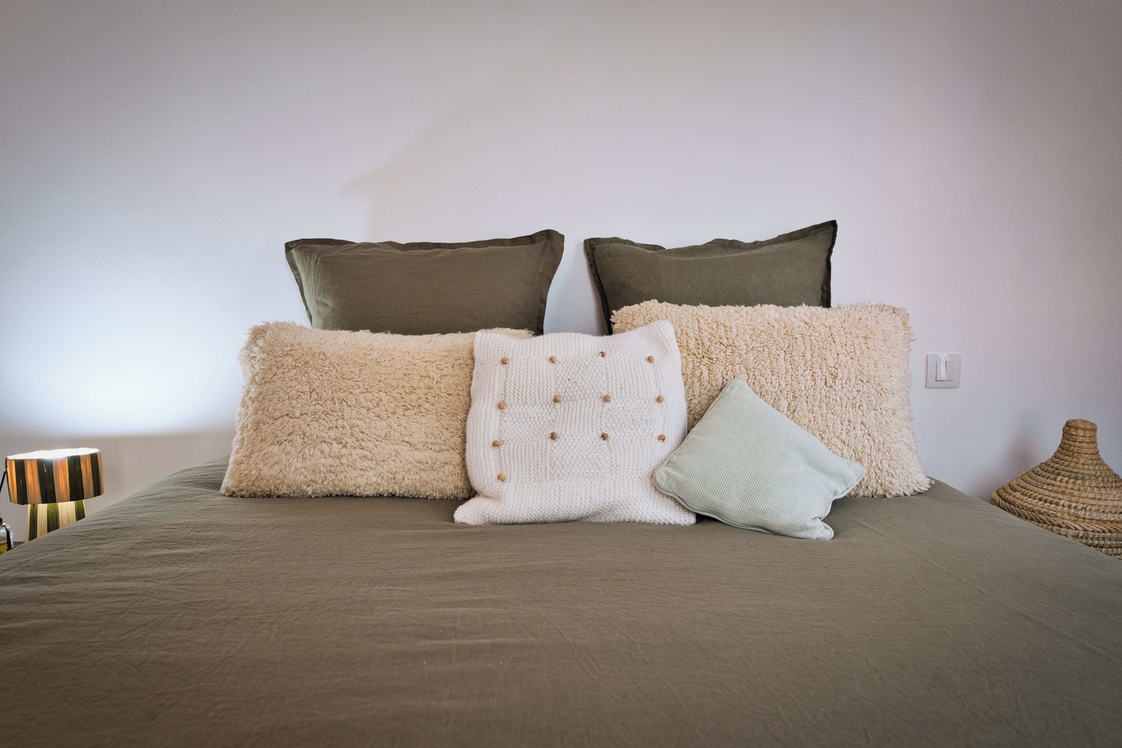 Una cama con una manta color beige y varias almohadas de distintas texturas y colores.