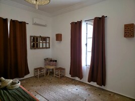 Una habitación con cama, ventana con cortinas, espejo, un taburete pequeño y una cesta.