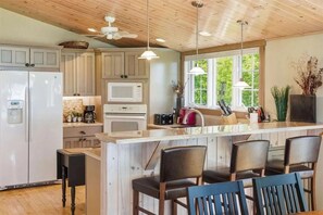 A kitchen with wooden cabinets, a refrigerator, a microwave, and a bar area with seating. - Lake Michigan Cottage - Sleeps 14 - Private Beach + Sunset Views (Harbor Springs)
