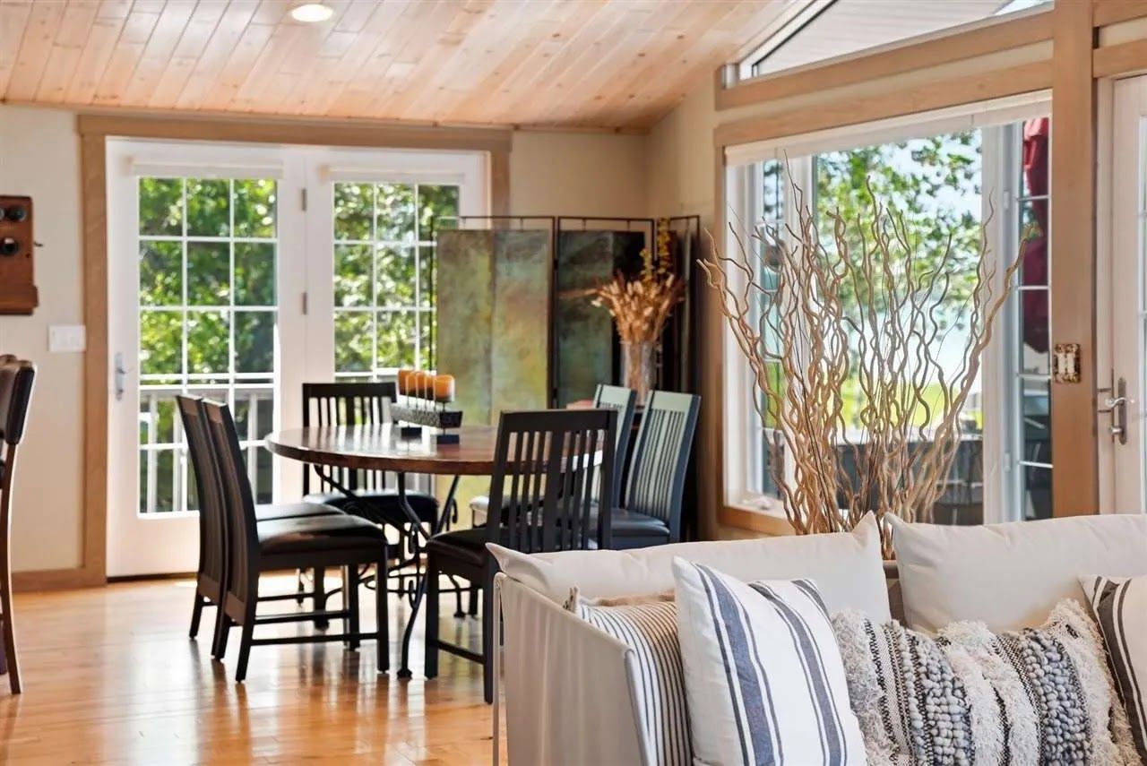 A dining area with a wooden table and chairs, a sofa with patterned cushions, and a large window with a view of greenery.