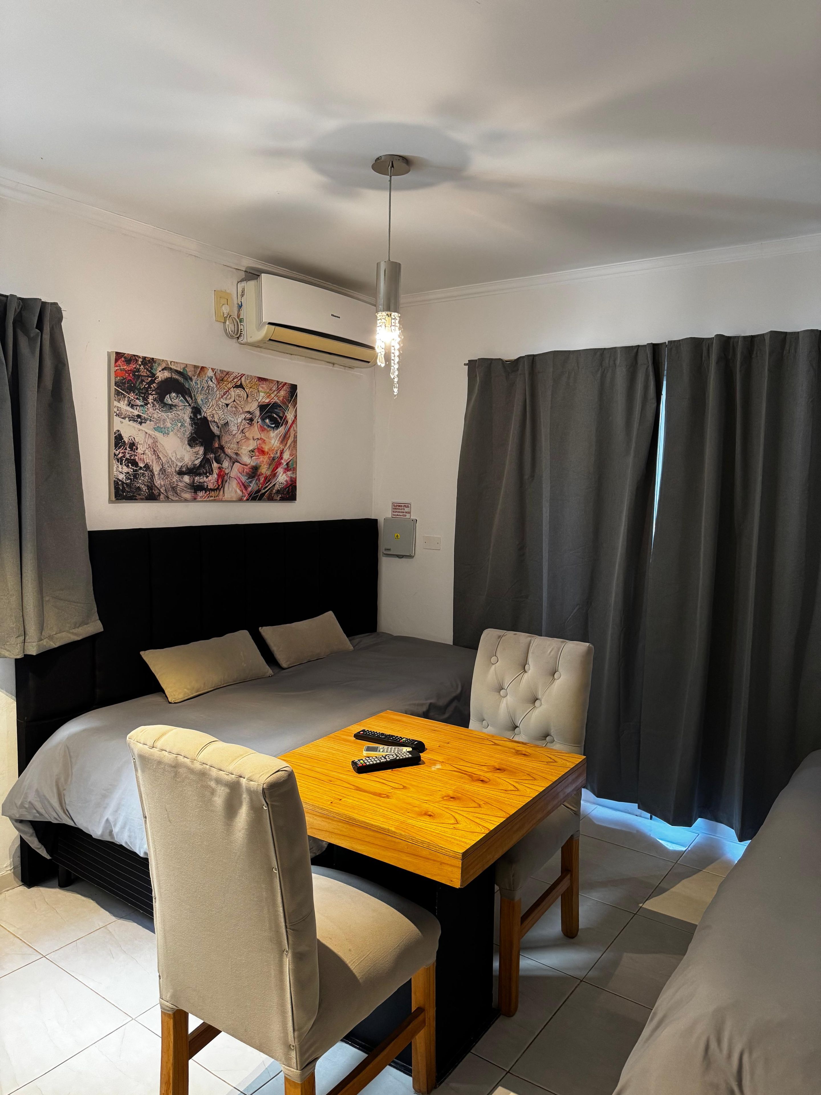 Habitación de hotel con cama, una mesa con sillas, equipo de aire acondicionado, un cuadro en la pared y cortinas.