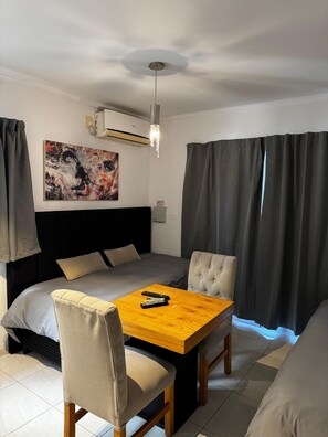 Habitació d'hotel amb un llit, una taula i cadires, un aparell d'aire condicionat, un quadre a la paret i cortines.