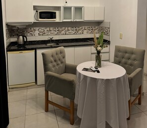 Una cocina compacta con microondas, tetera y lavamanos. Hay una mesa redonda con sillas y un jarrón con flores.