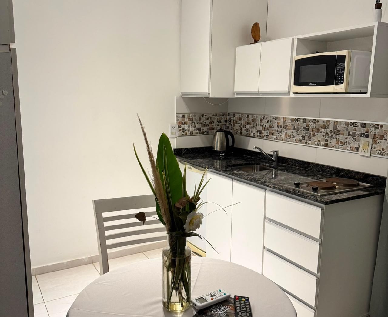 Una pequeña cocina con armarios blancos, microondas, cafetera y un florero con flores sobre una mesa redonda.