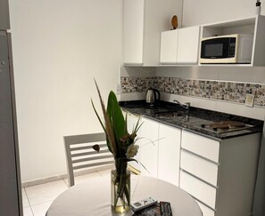 Una cuina petita amb armaris blancs, microones, cafetera i un ram de flors sobre una taula rodona.