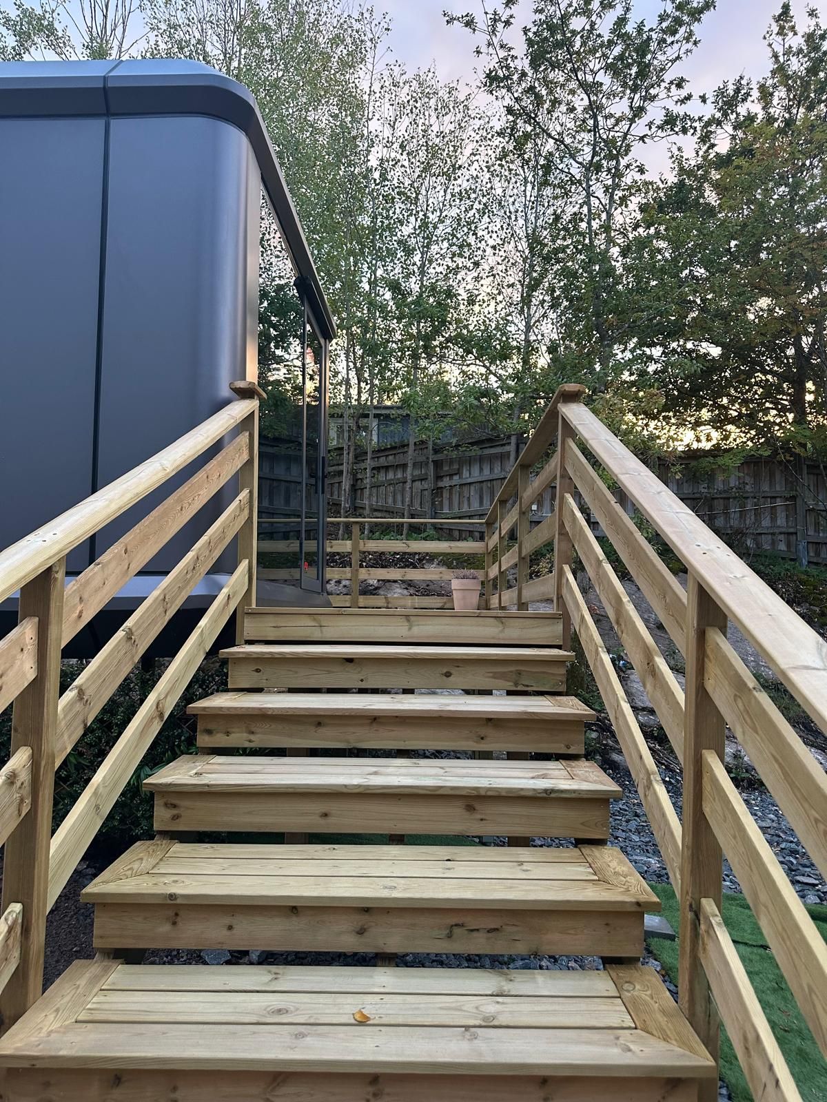 A wooden staircase leading to a deck area with a view of trees and a fence in the background.