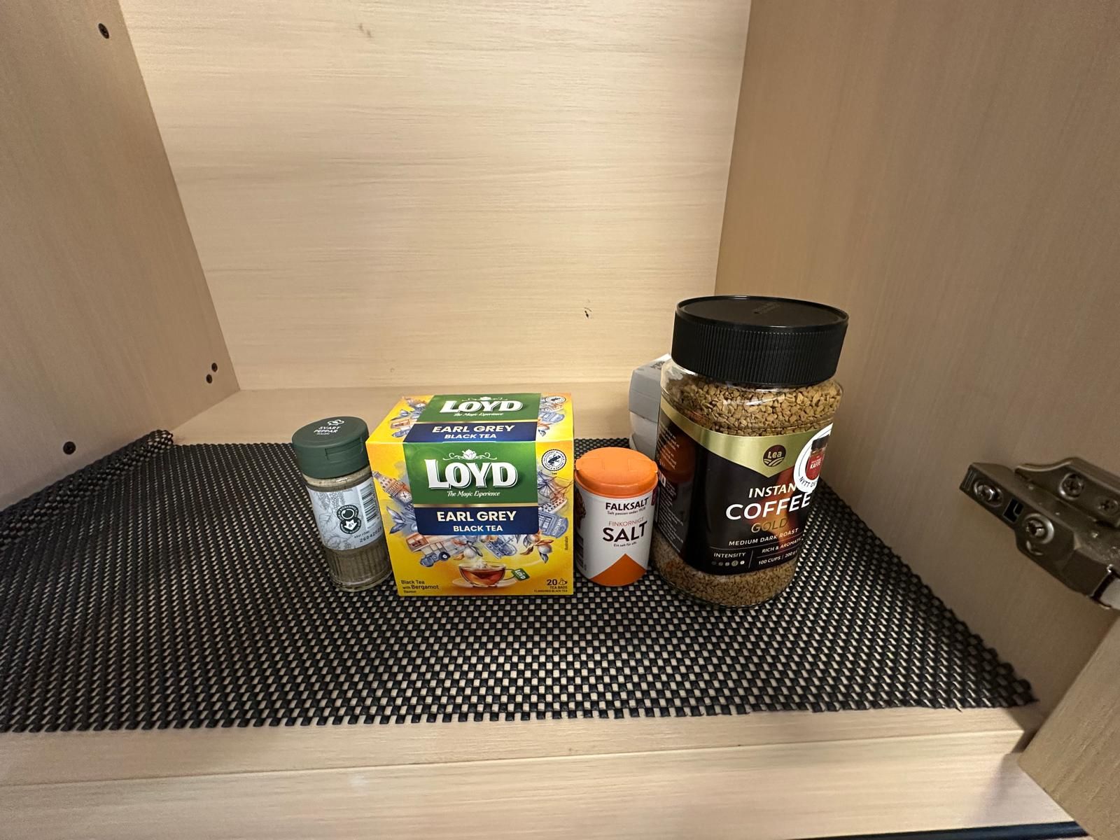 A drawer with a black mesh mat, containing a box of Earl Grey black tea, a salt container, and a jar of instant coffee.