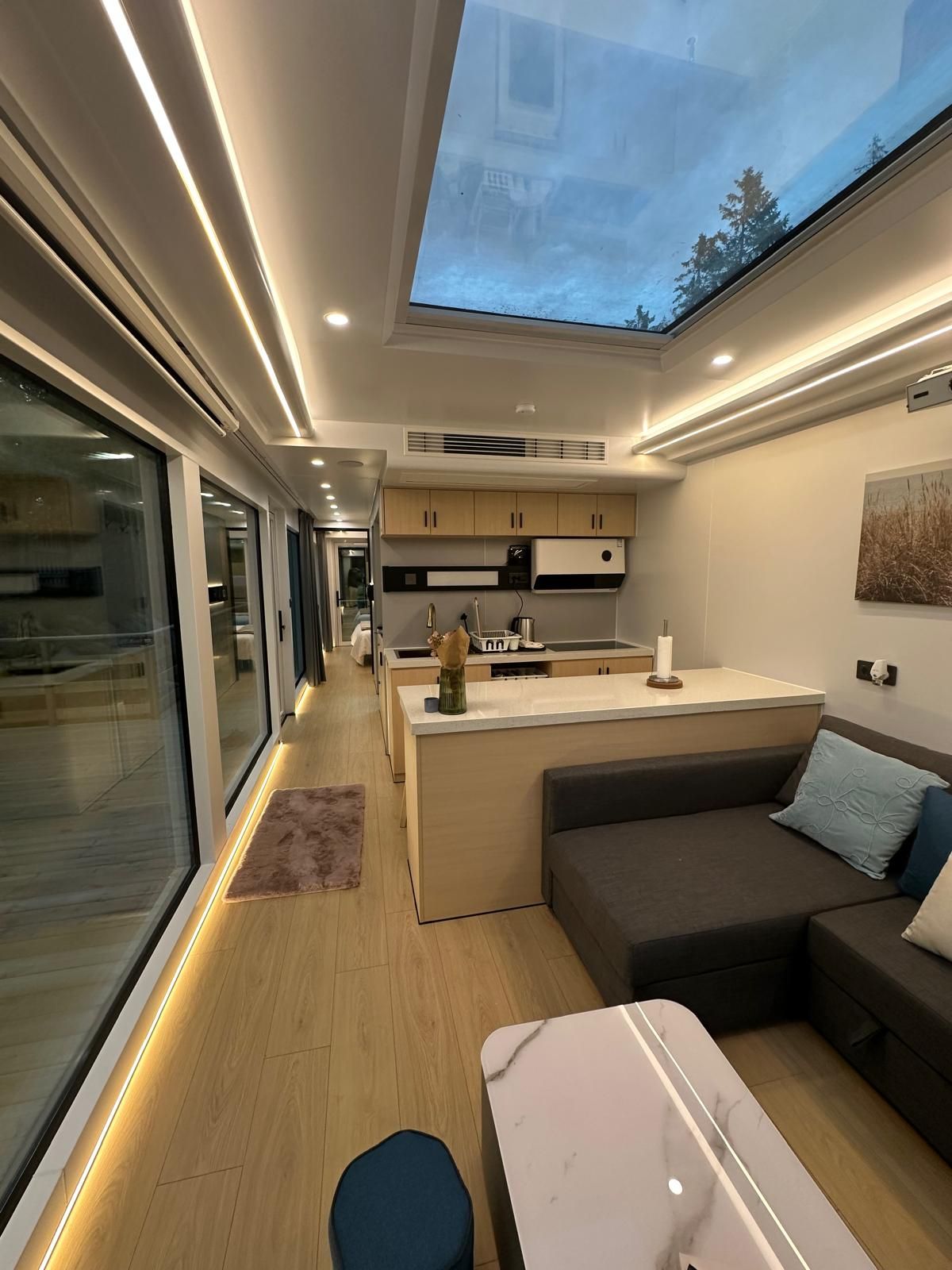 A modern interior with a kitchen, dining area, and seating space, featuring a skylight and wooden flooring.