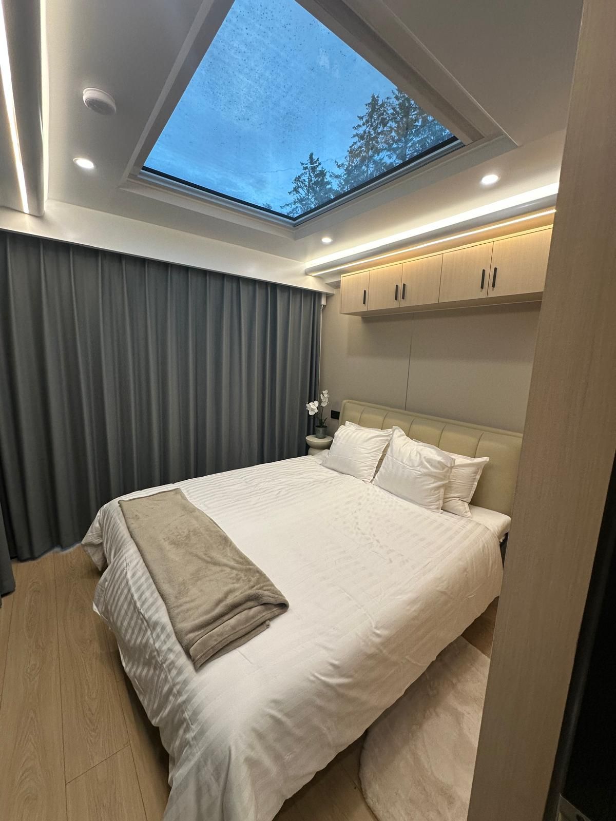 A bedroom with a skylight, a bed with white linens, a nightstand, and a view of trees through the window.