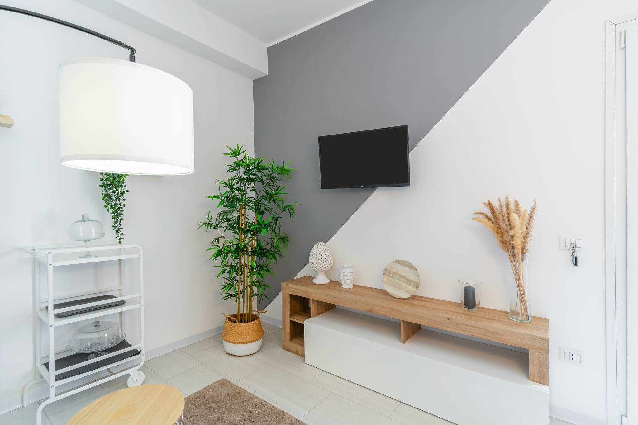 A modern living room with a white console table, a large potted bamboo plant, a flat-screen TV mounted on the wall, and a wooden bench with decorative items.