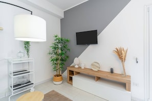 Ein modernes Wohnzimmer mit einem weißen Konsolentisch, einer großen Kübelpflanze mit Bambus, einem flachbildfernseher an der Wand und einer Holzbank mit Dekorationselementen.
