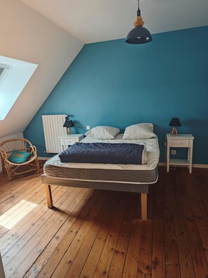 A bedroom with a sloped ceiling, a bed with a dark blanket, a white bedside table with a lamp, a wicker chair, and a window. - Meurdrac's Rest (La Meurdraquière)