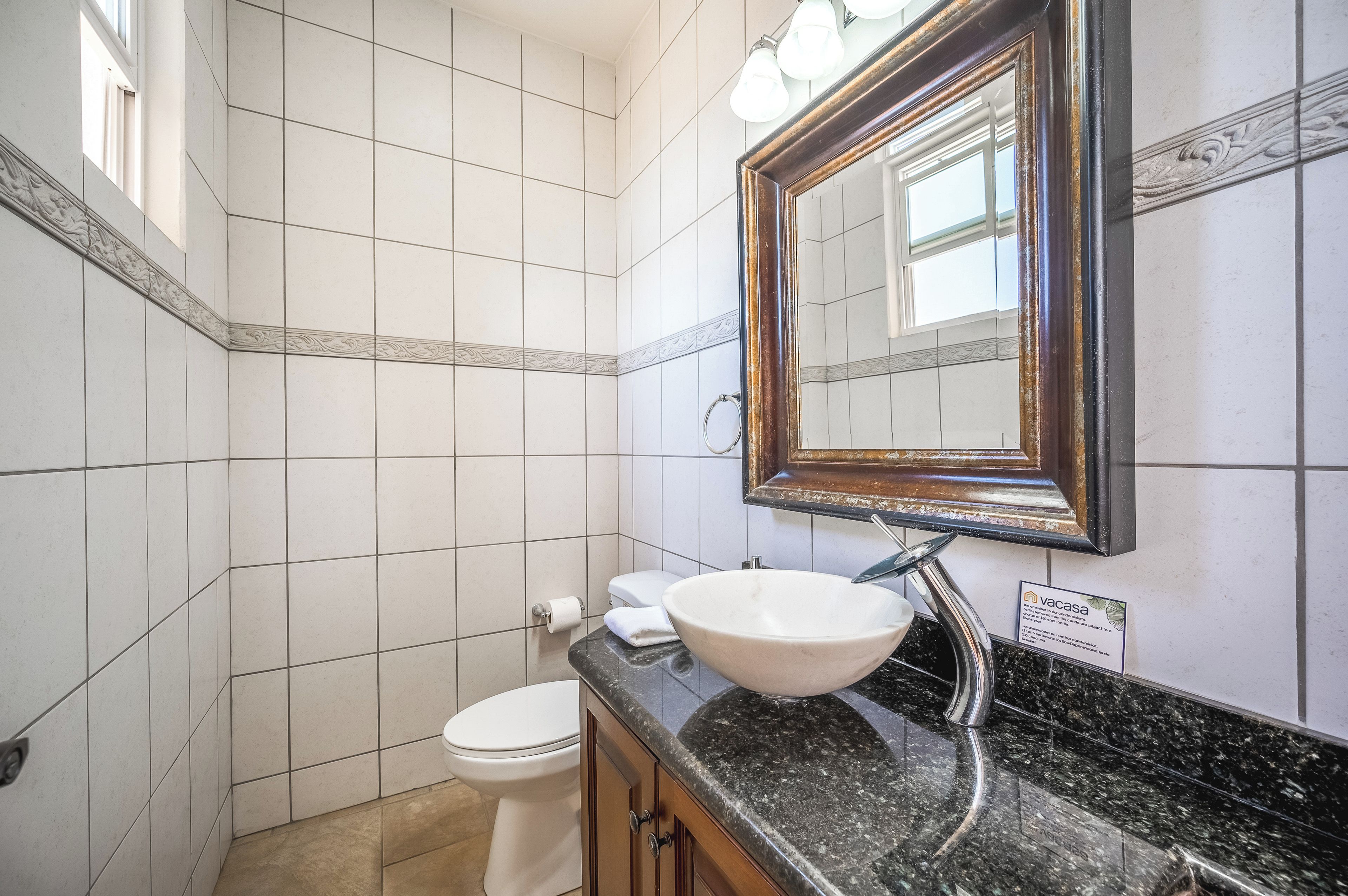 A bathroom with white tiled walls, a white toilet, a sink with a marble countertop, and a mirror with a decorative frame.