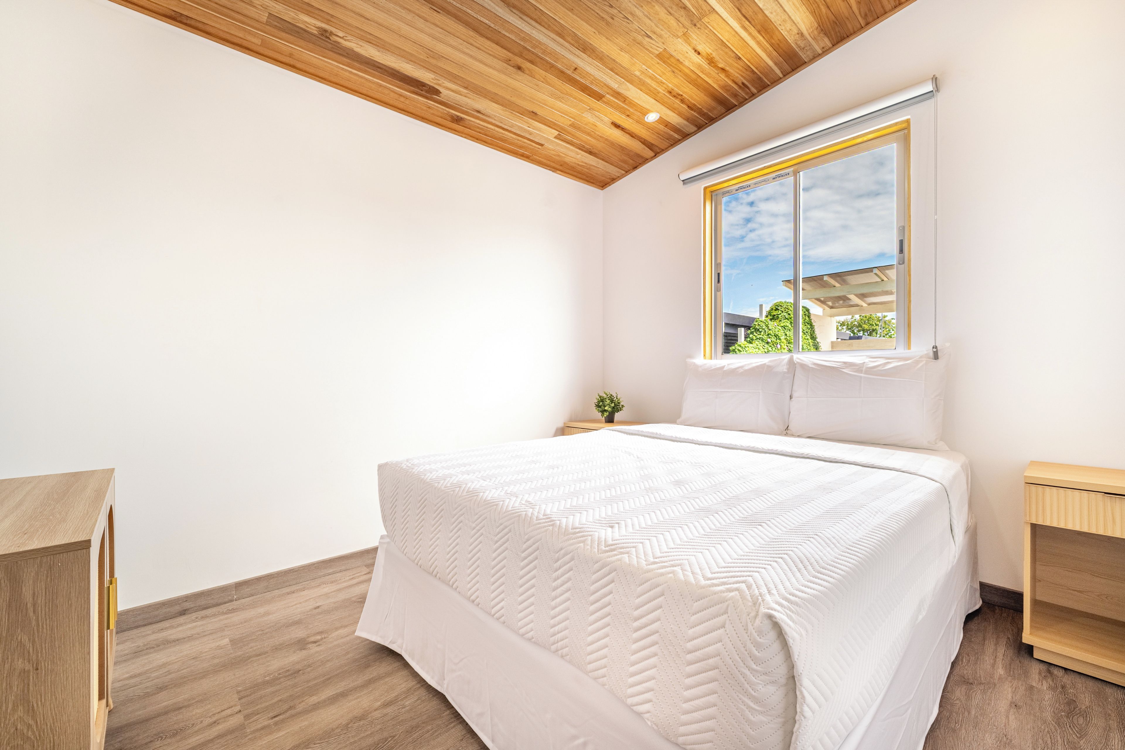 A bedroom with a wooden ceiling, a large bed with a white quilt, a window with a view of greenery, and a bedside table with a plant.