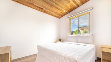 A bedroom with a wooden ceiling, a large bed with a white quilt, a window with a view of greenery, and a bedside table with a plant.