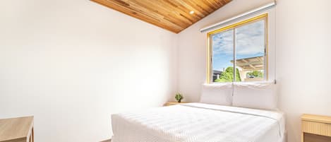 Un dormitorio con techo de madera, una cama grande con colcha blanca, una ventana con vistas a la vegetación y una mesita de noche con una planta.