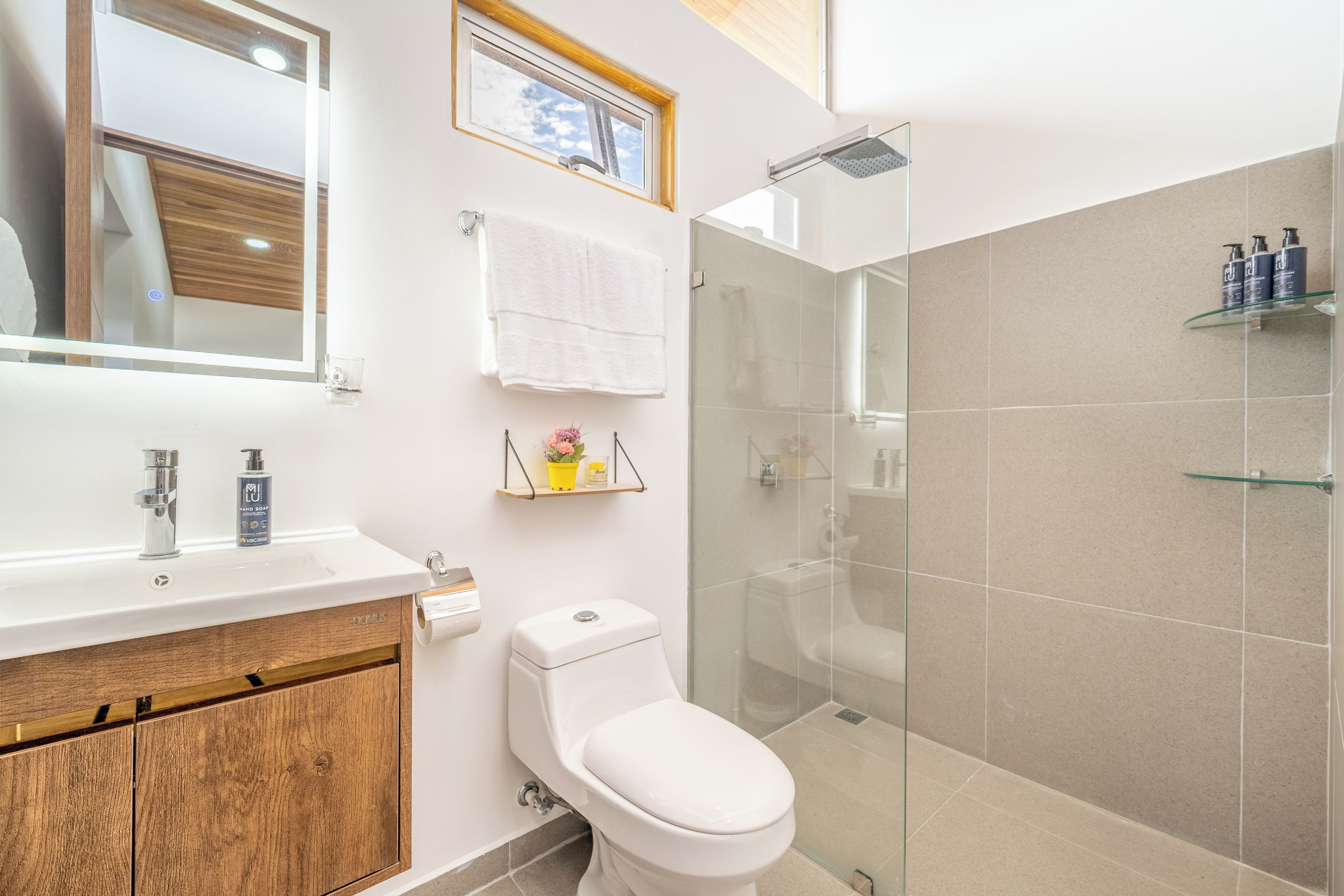 Une salle de bain moderne avec un meuble-lavabo en bois, des toilettes, une douche en verre et une étagère contenant des articles de toilette.