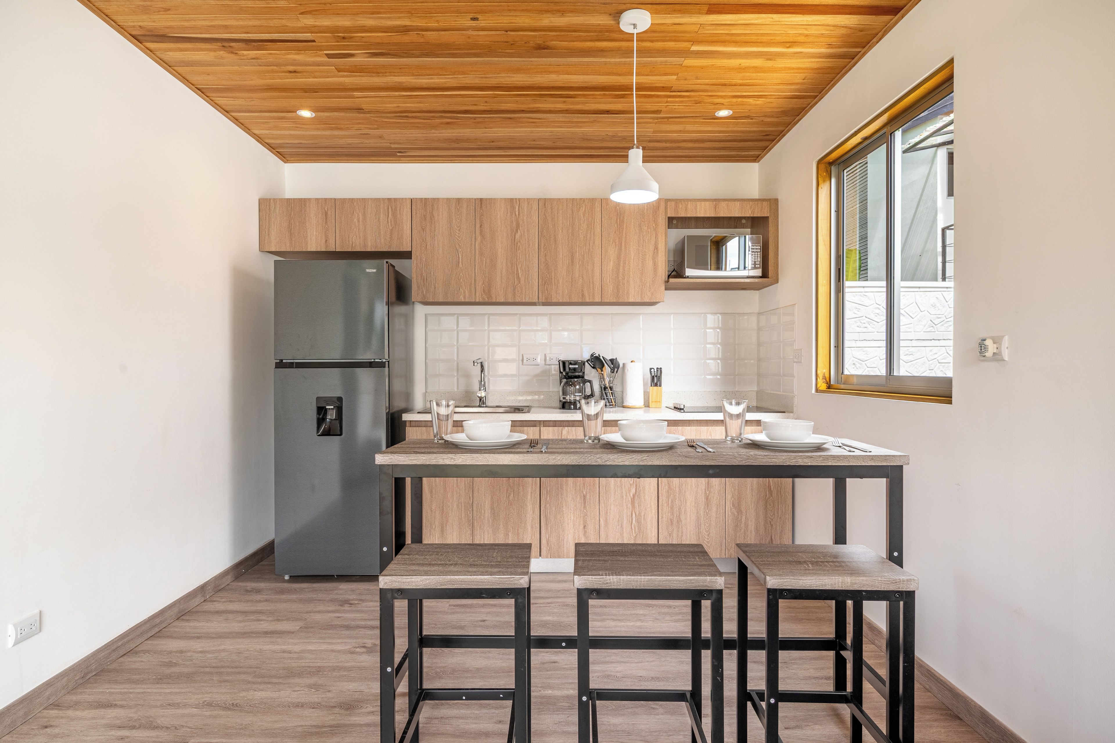 A modern kitchen with wooden cabinets, a stainless steel refrigerator, a central island with bar stools, and a pendant light.