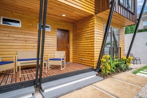A wooden exterior with a door marked '6', a covered patio with wicker furniture, and a small garden with yellow flowers.