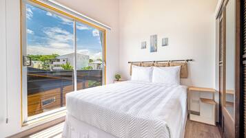 A bedroom with a large window, a bed with white bedding, a bedside table, and a nightstand.