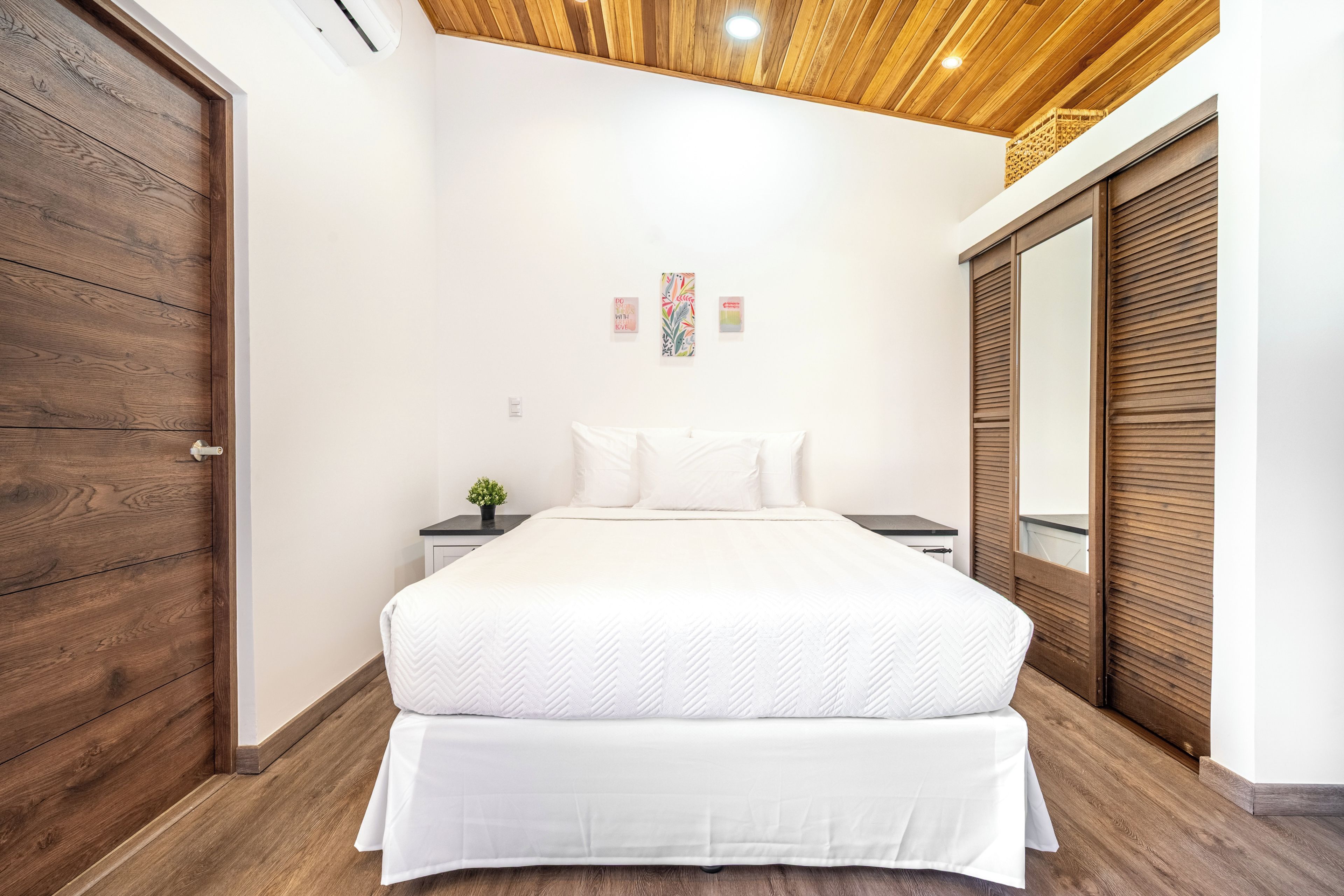 A bedroom with a wooden ceiling, a bed with white bedding, a nightstand with a plant, and a wardrobe with a mirror.