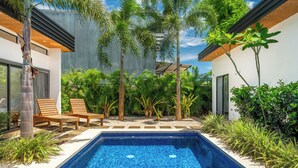 Piscina exterior moderna com borda em azulejo, rodeada por vegetação exuberante e um deck de madeira com espreguiçadeiras.