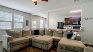 A living room with a sectional sofa, a kitchen area with dark cabinets, and a 'Hello' sign on the wall.