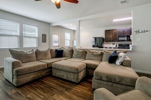A living room with a sectional sofa, a kitchen area with dark cabinets, and a 'Hello' sign on the wall.