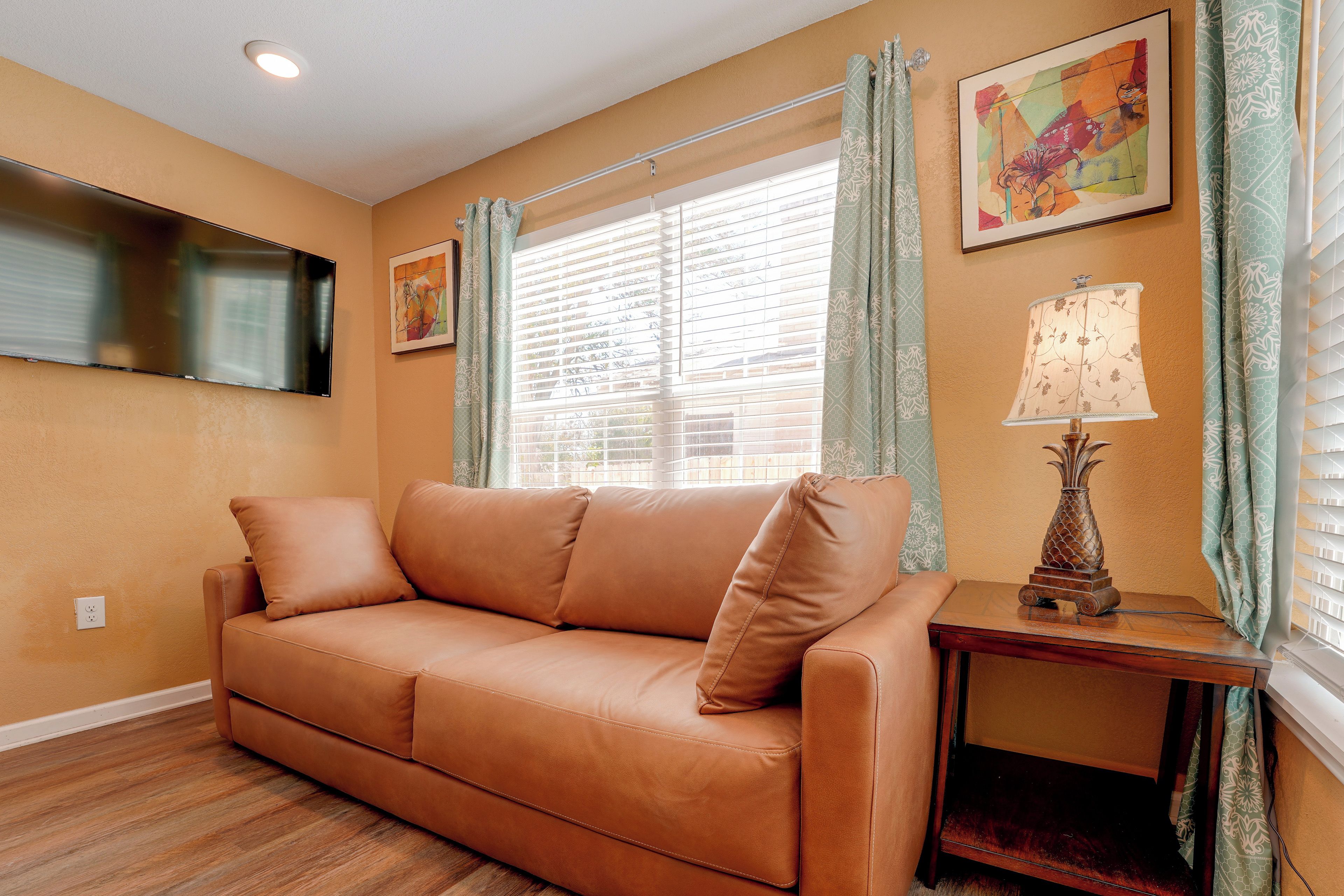 A tan leather sofa with a wooden side table and a lamp, positioned in front of a window with blinds.