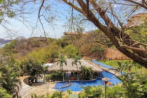 Área de piscina com estrutura de telhado de palha, cercada por palmeiras e vista para uma colina com edifícios.
