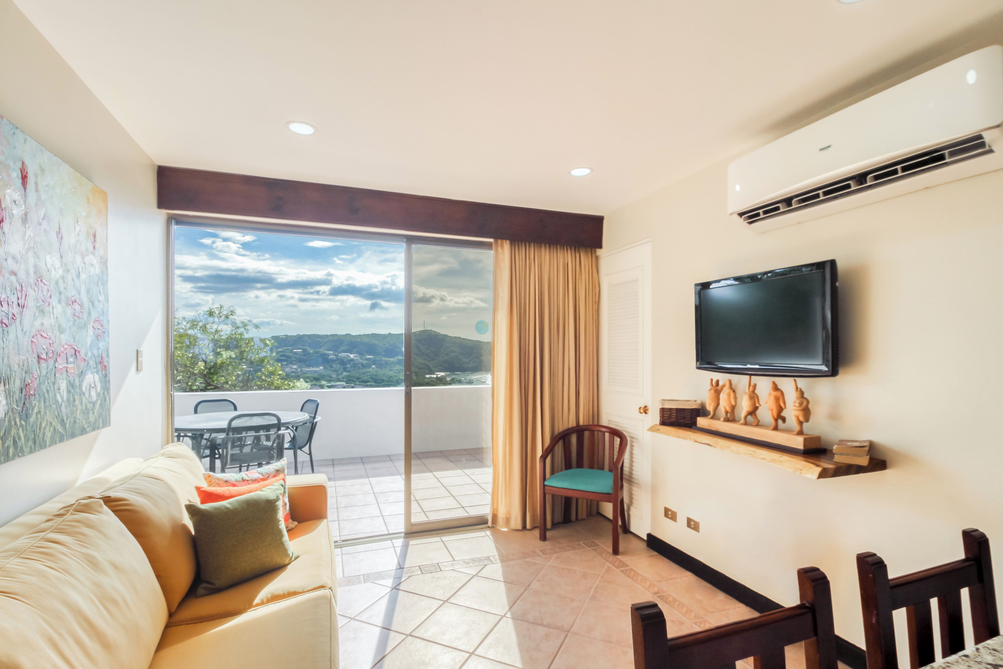 A living room with a sofa, a TV mounted on the wall, a dining area with chairs, and a sliding glass door leading to a balcony.