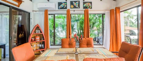 Comedor con una mesa dispuesta para cuatro personas, sillas naranjas, un centro de mesa decorativo y vistas al exterior con vegetación.