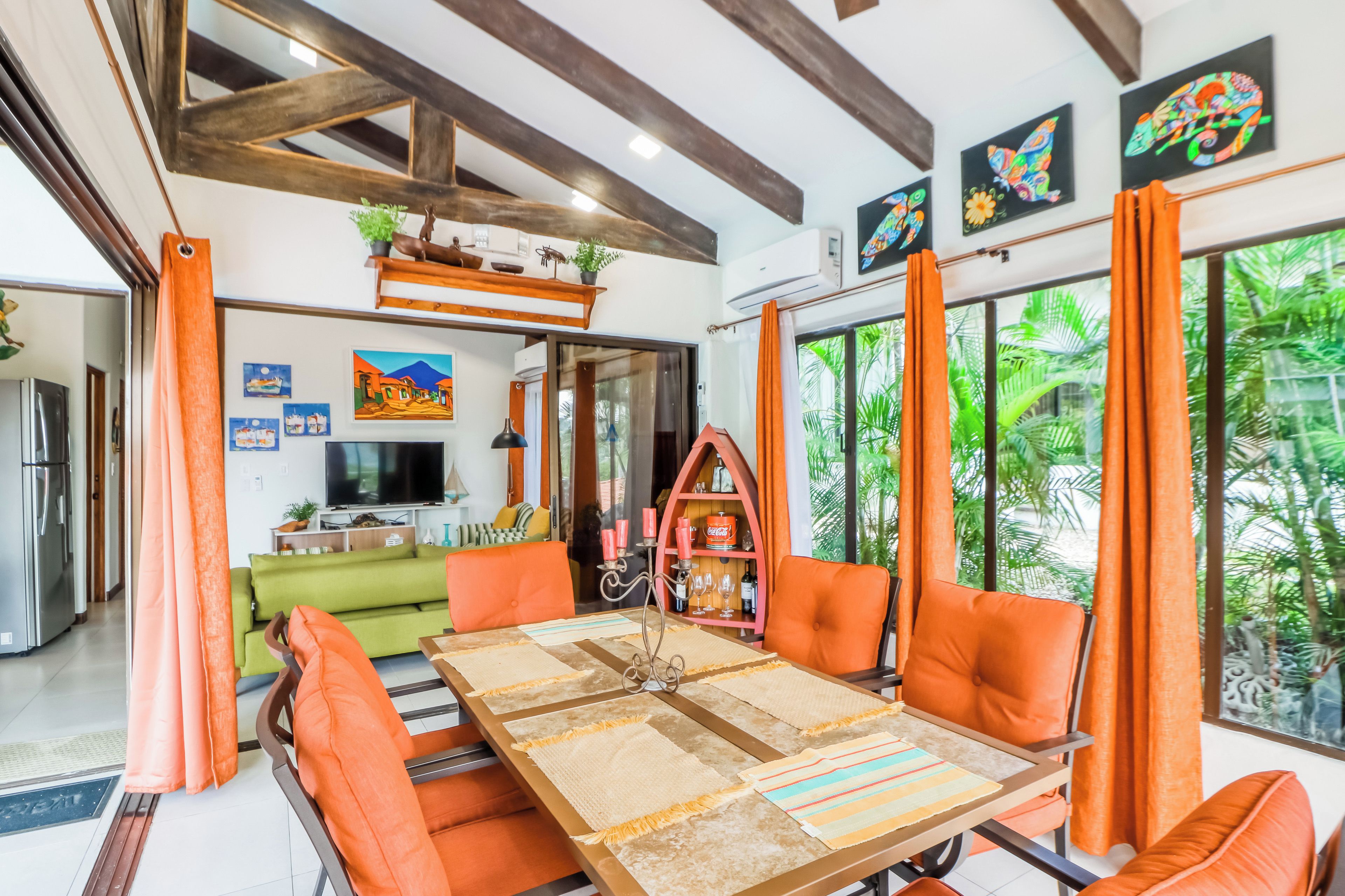 Un salon moderne doté d’un grand téléviseur, d’un coin repas avec des chaises orange et offrant une vue sur une végétation tropicale luxuriante par les fenêtres.