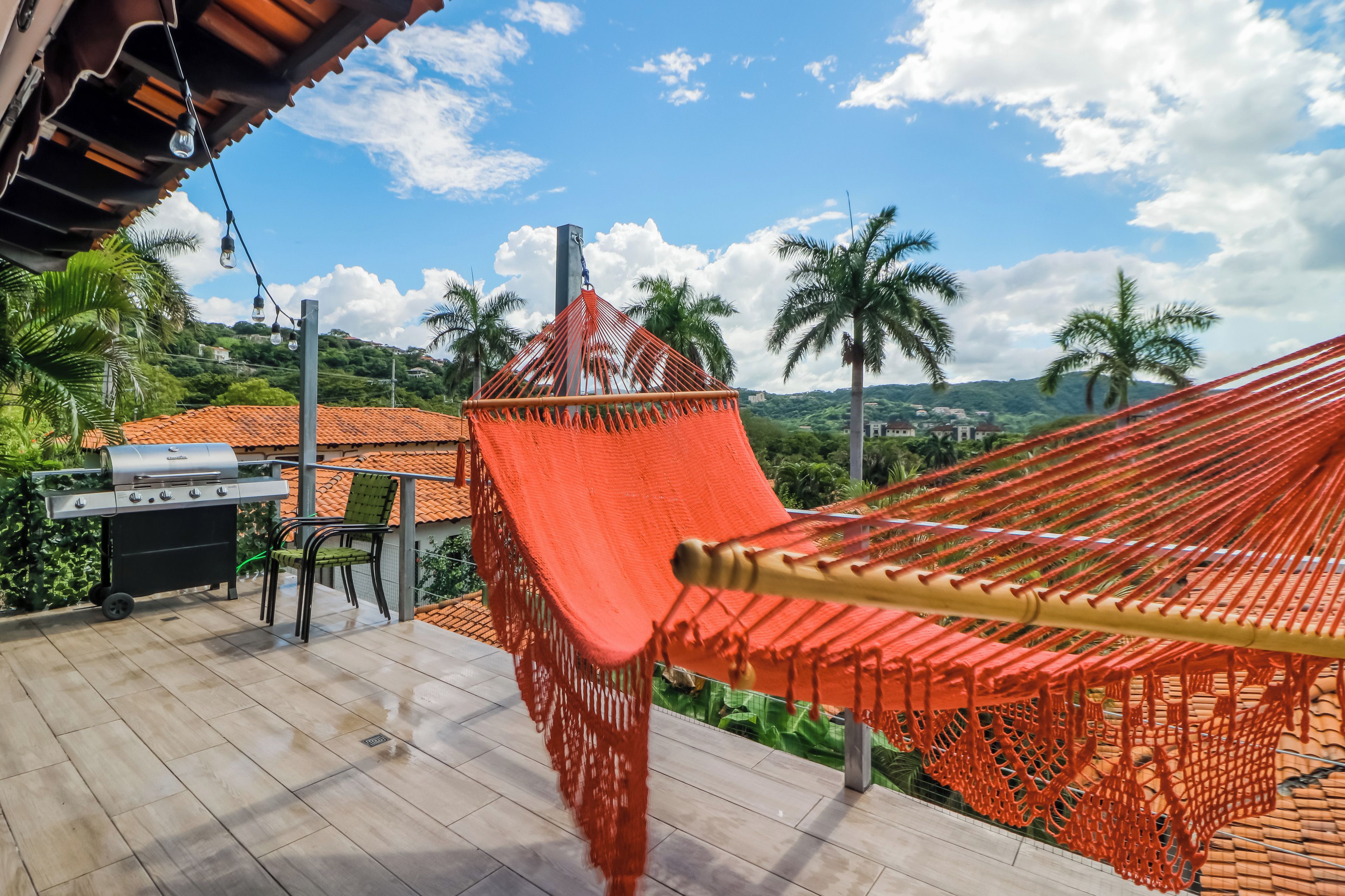 Una hamaca sobre una terraza de madera con una parrilla y una silla, con vistas a un paisaje de palmeras y edificios.