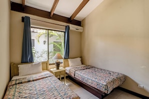Quarto com duas camas, janela com cortinas, ar condicionado e abajur em uma mesinha de cabeceira.