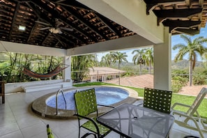 Área externa coberta com piscina, assentos e vista para palmeiras e uma casa.