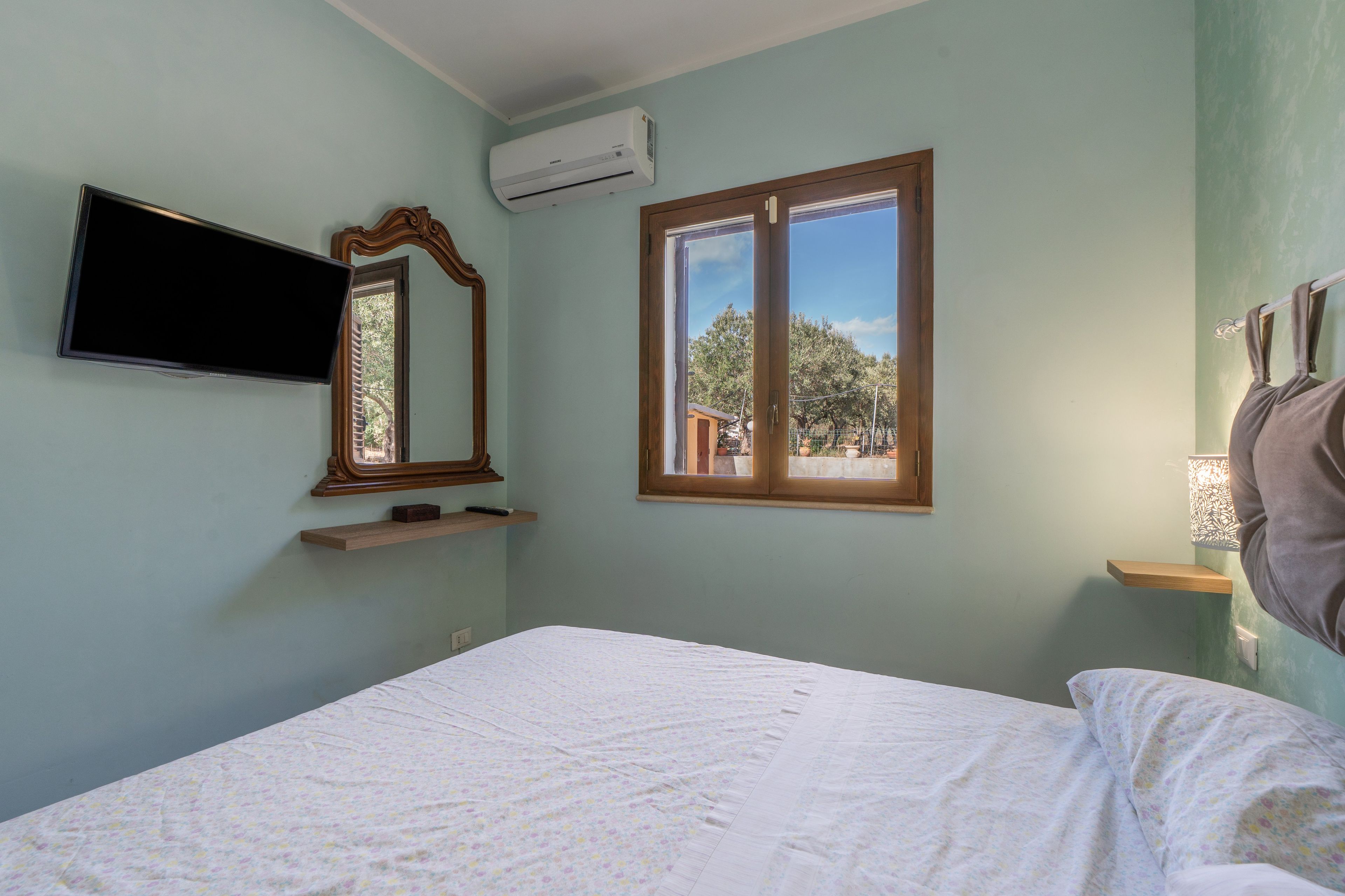 A bedroom with a bed, a mounted TV, an air conditioner, a window, and a mirror.