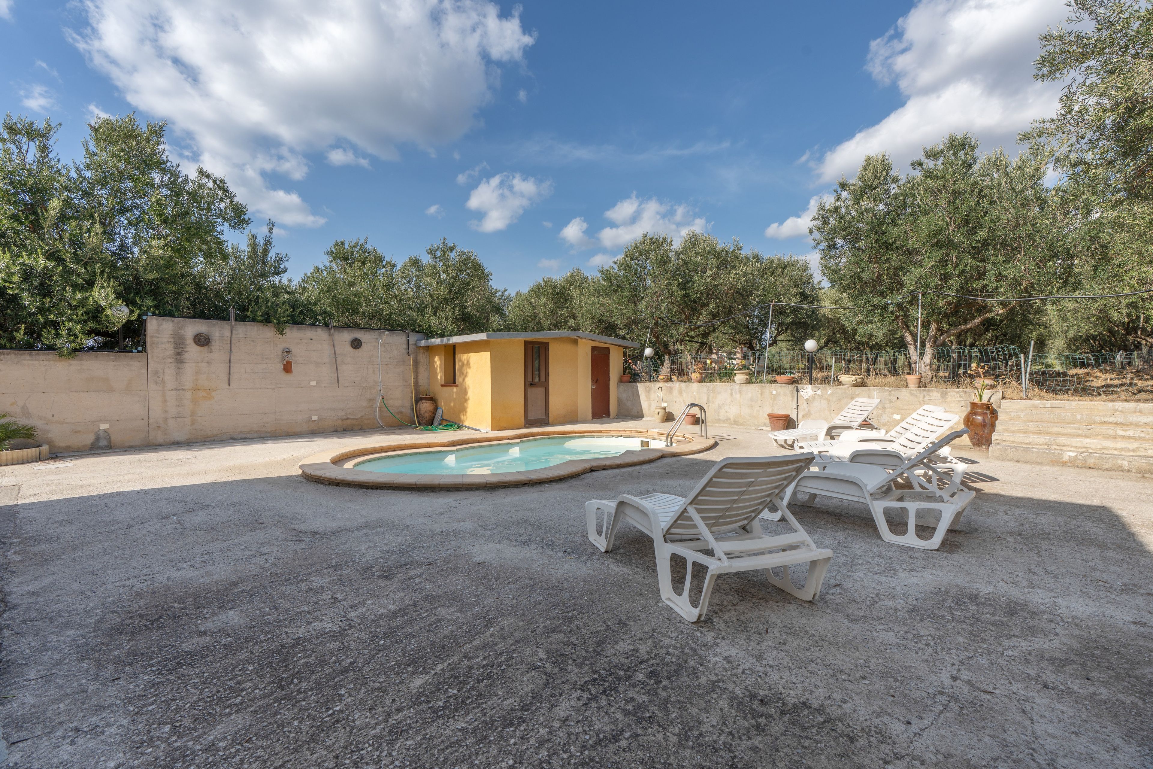 A pool area with lounge chairs and a small building, surrounded by trees and a concrete wall.