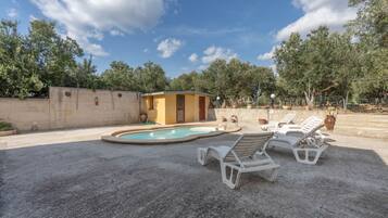 A pool area with lounge chairs and a small building, surrounded by trees and a concrete wall.
