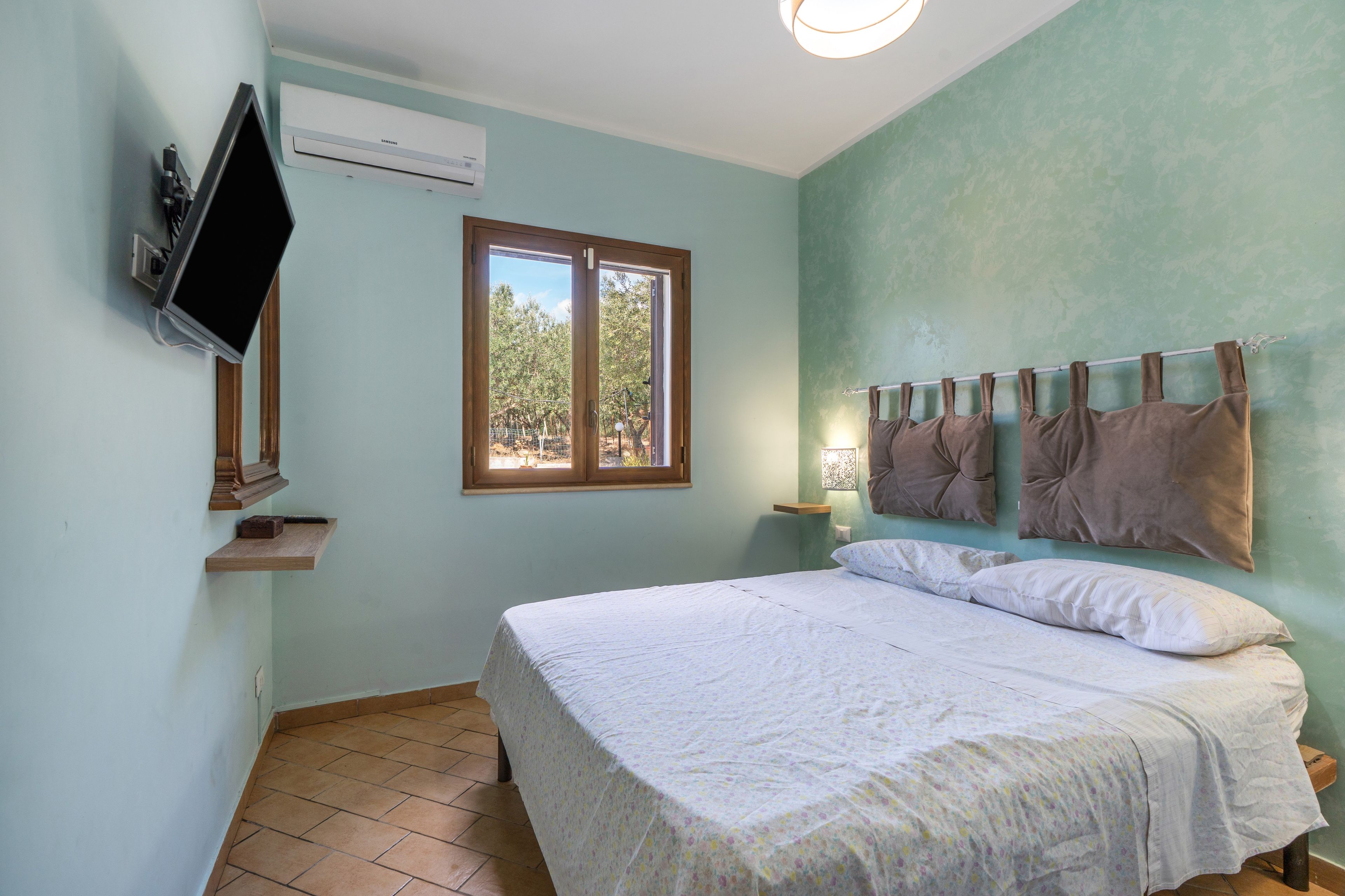 Une chambre à coucher avec un lit, un téléviseur fixé au mur, une fenêtre et une table de chevet avec une lampe.