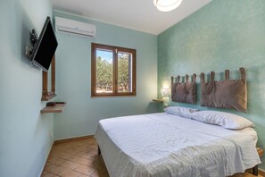 A bedroom with a bed, a TV mounted on the wall, a window, and a nightstand with a lamp.